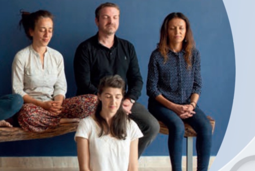 A group of four people all wearing shades of blue, practicing meditation in front a blue-painted wall.