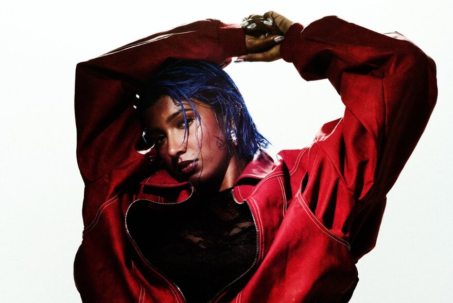 Singer Charlotte Dirckze posing for the camera against a white background, wearing an oversize red jacket, her arms raised and resting on her head.