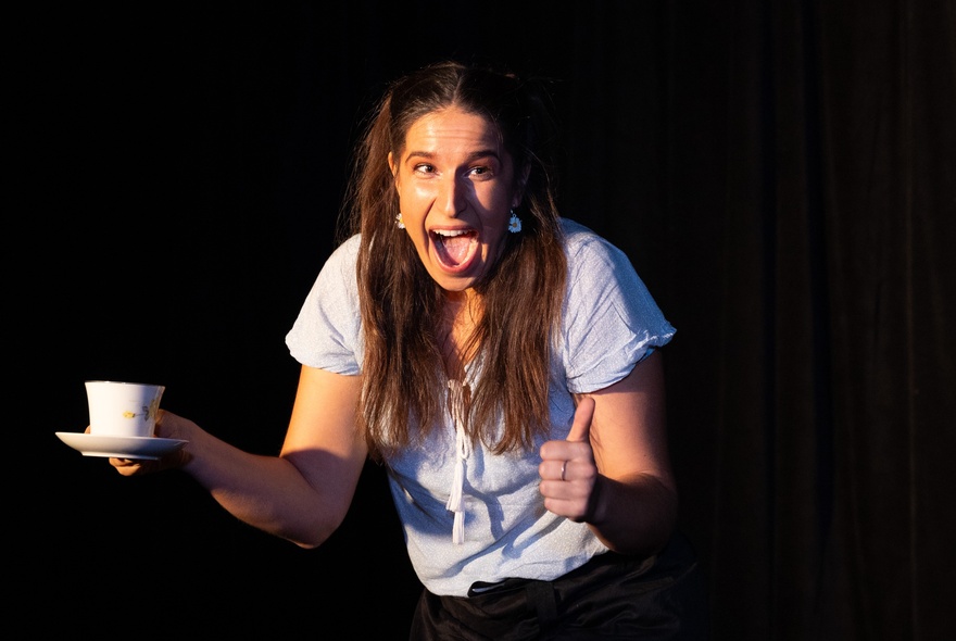 A performer on a dark stage with a teacup and saucer during a show, her mouth open wide.
