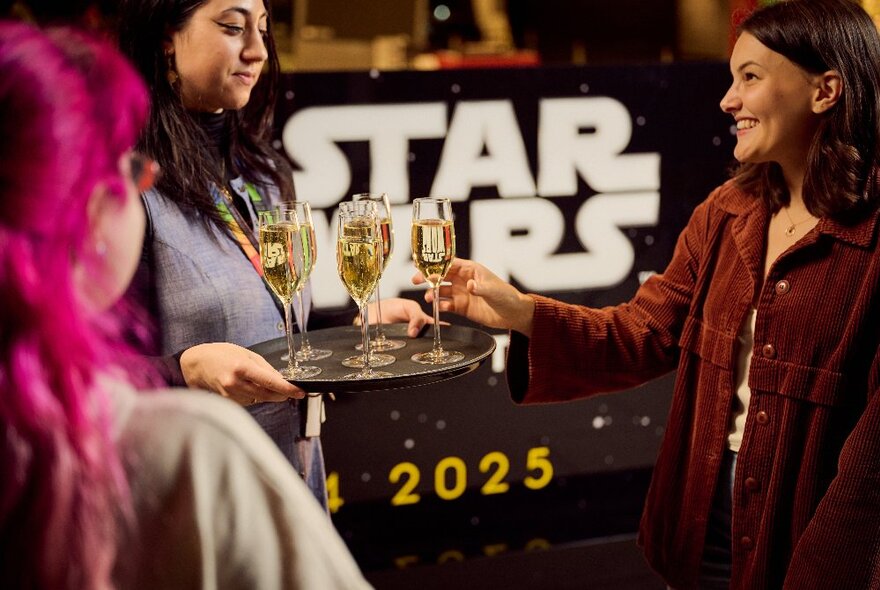 A person taking a glass of champagne from a tray from a waiter with Star Wars signs in the background. 