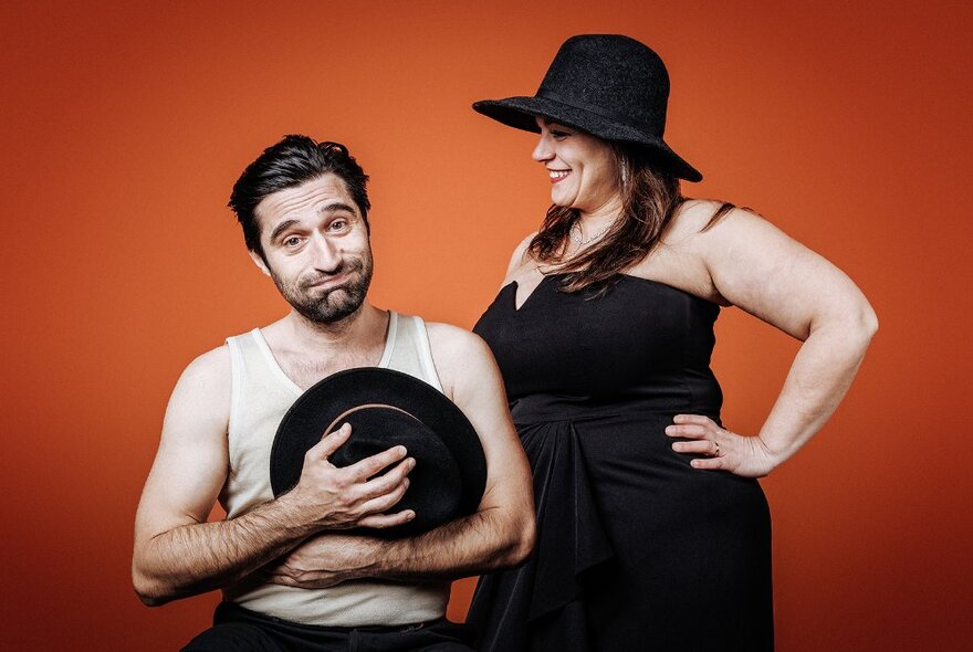 A seated man wearing a white singlet and holding a black hat to his chest looking bemused, with a smiling woman standing next to him in a black dress and black hat.