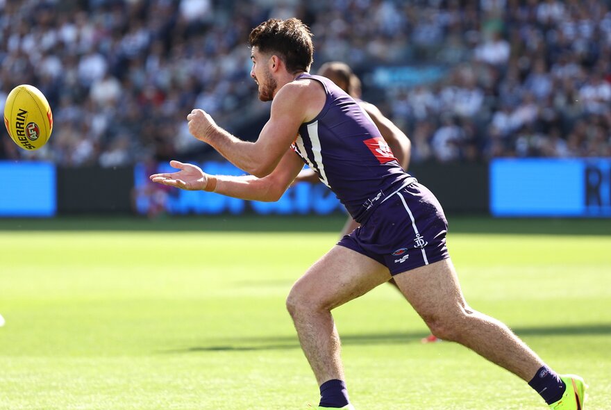 A Fremantle AFL football player running on the field during a match, a blurred crowd behind him.