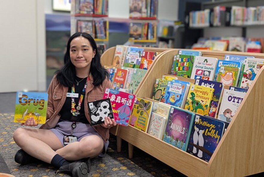 A smiling library worker holding up three picture books with their front covers facing out, seated cross-legged on the floor in front of library display shelves.