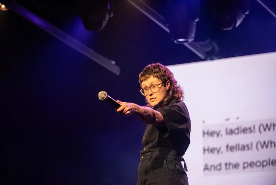 Comedian performing on stage holding a microphone in front of a screen.