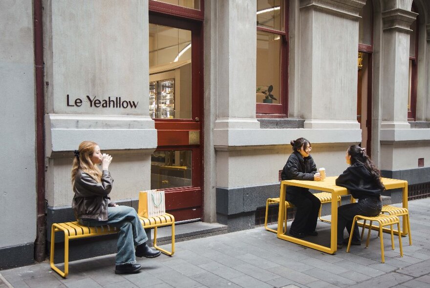 The exterior of the cafe and cake shop Le Yeahllow, with people sitting outside on yellow benches and chairs on the city footpath, eating and drinking.