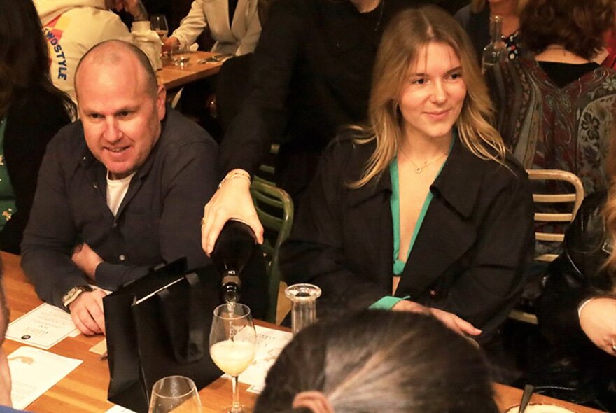 Two people seated at a table in a restaurant, glasses of wine in front of them, conversing with other people seated at the table.