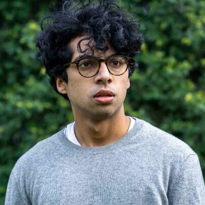 Comedian Rao looking anxious, with dark hair and glasses, against tree backdrop.