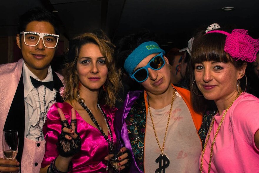 People dressed in 1980s outfits, neon t-shirts and large sunglasses smiling for the camera on a boat cruise.