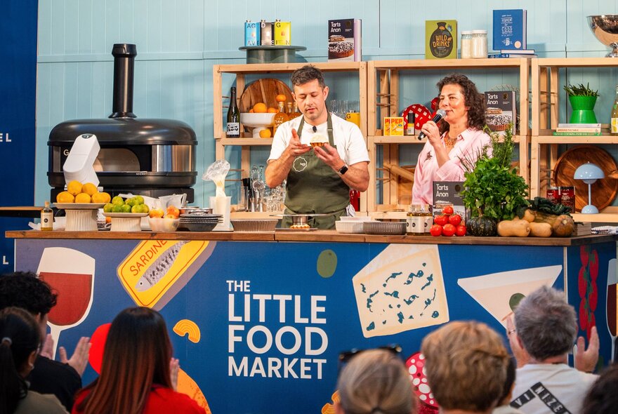 A live cooking and tasting demonstration at The Little Food Market, featuring a chefs and an MC on stage in front of an audience.