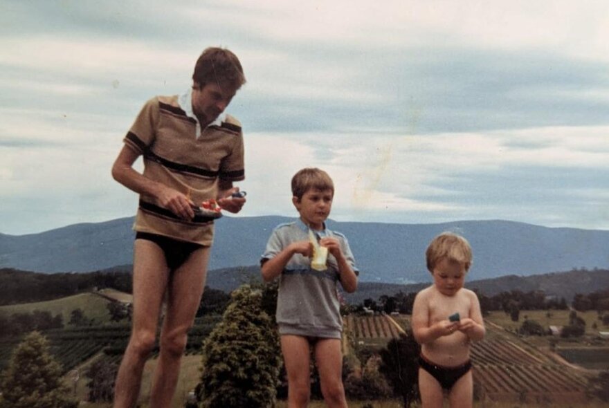 A vintage candid photograph featuring two young boys outdoors in swimwear next to their father, against a scenic hilly landscape backdrop.