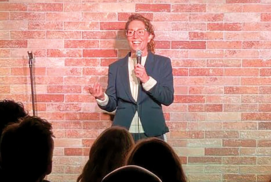 Comedian Kitty Flanagan performing stand-up at a small venue with a brick wall background. 