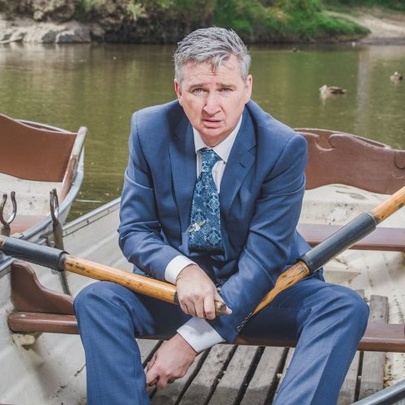 Comedian Damian Callinan wearing a navy suit, shirt and tie, sitting in a row boat in a river, holding an oar in each hand.