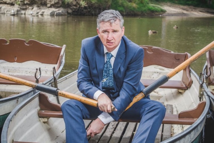Comedian Damian Callinan wearing a navy suit, shirt and tie, sitting in a row boat in a river, holding an oar in each hand.