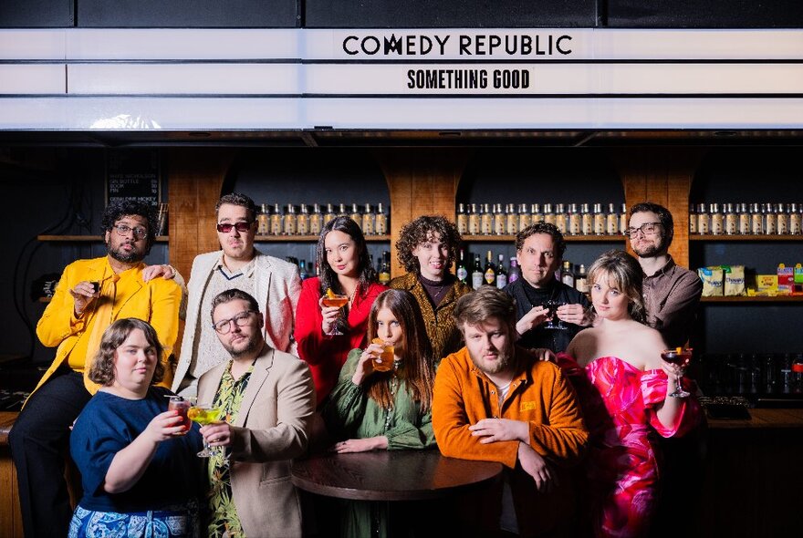 A group of 11 performers arranged around at table at a bar that says Comedy Republic Something Good. 