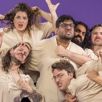 A group of actors dressed in beige clowning around for a group photo. 