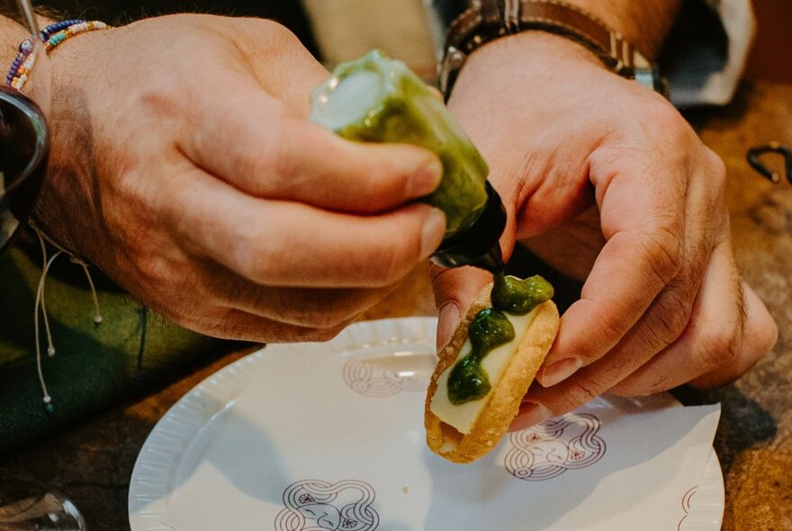 Hands holding a small snack and squeezing green sauce on it. 