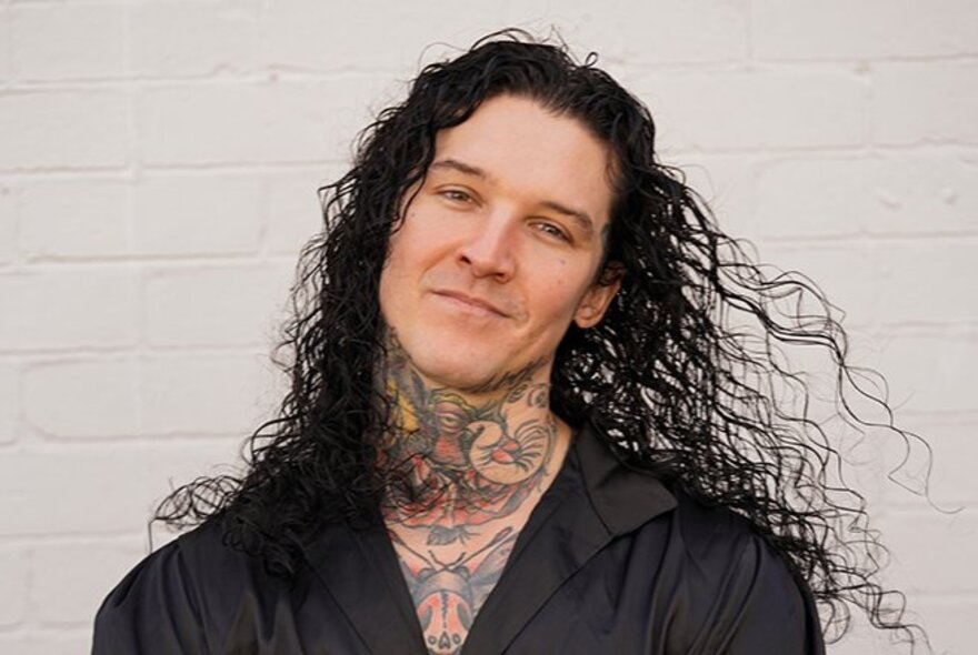 A man with long, dark, curly hair and extensive neck and chest tattoos, smiles slightly while posing against a white brick wall.