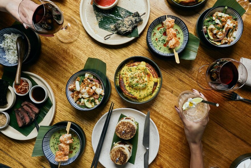 Looking down at dishes of food on a wooden dining table, with cutlery and hands holding glasses also visible.