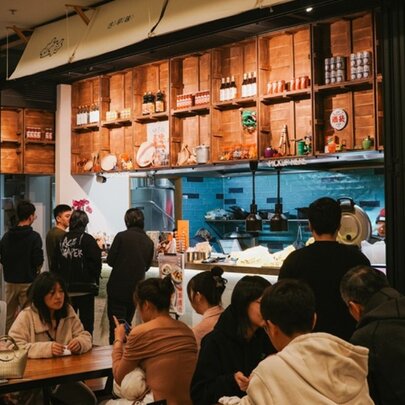 Diners at tables and ordering counter in a busy Asian cafe with wooden shelving details.