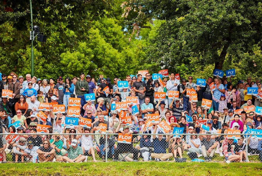 People seated and standing on the side of a grassy river bank, holding up orange signs that say FLOP or blue signs that say FLY, watching the Birdman Rally in progress.