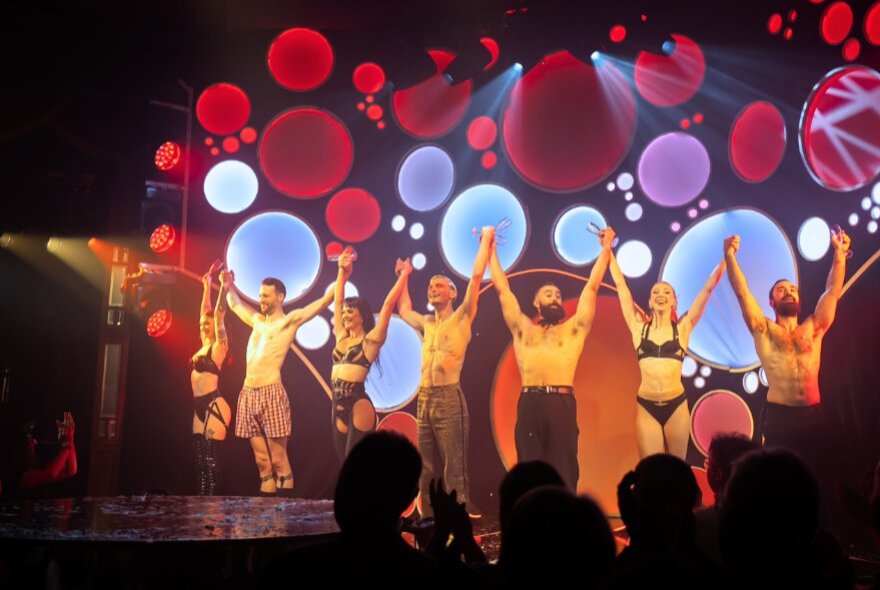 An ensemble of cabaret performers standing in a line on stage with their arms raised and holding hands, about to take a bow, the audience before them clapping and cheering.
