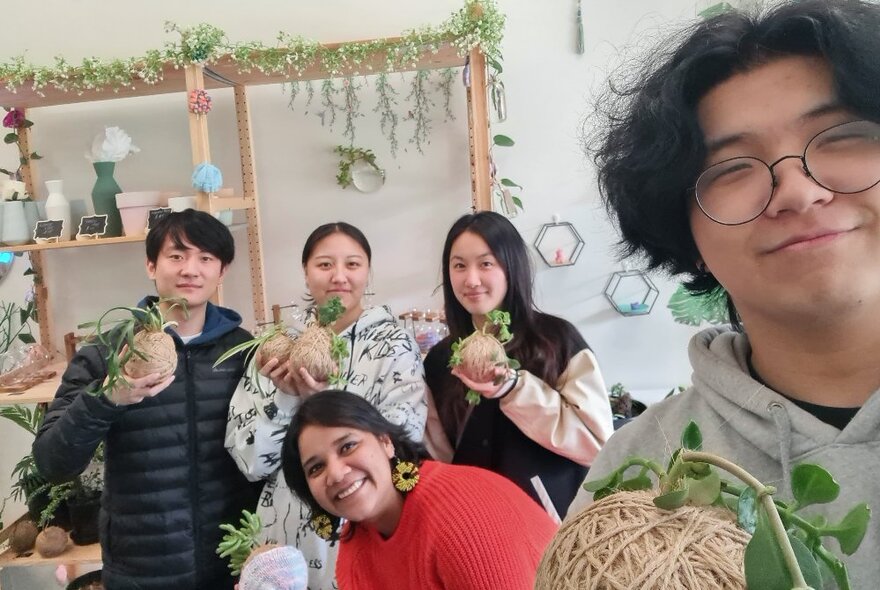 Class participants holding up their home-made kokedama balls of string topped with plants, in a workshop room with shelves draped with plants and crafts.