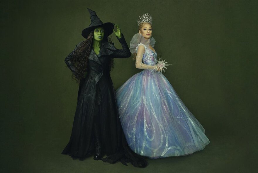 Models dressed in elaborate costumes for the characters of Elphaba, with green skin and wearing a black gown and pointed hat and Glinda, wearing a shimmering white gown and tiara, from the Wicked films.