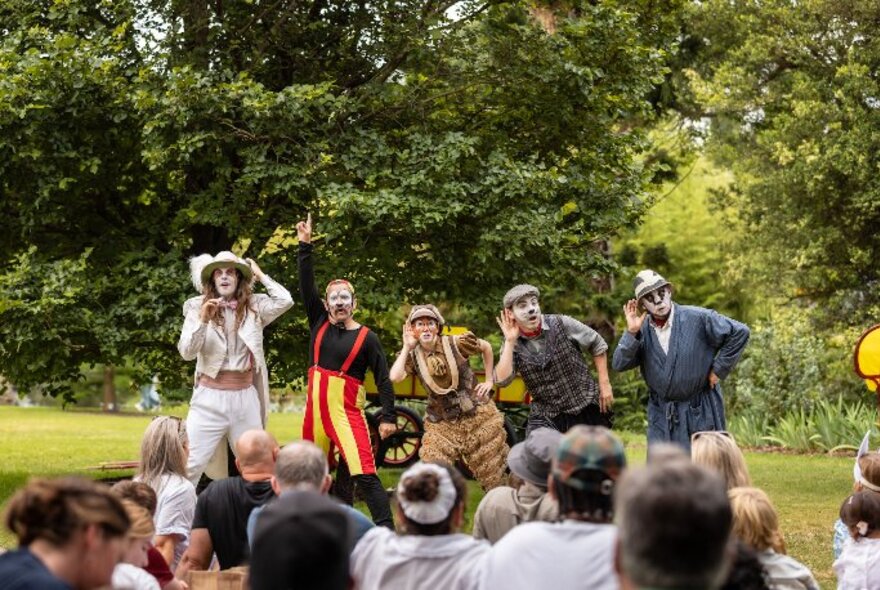 Costumed actors dressed as Wind in the Willows characters performing against a backdrop of trees and grass, with an audience seated on the grass before them.