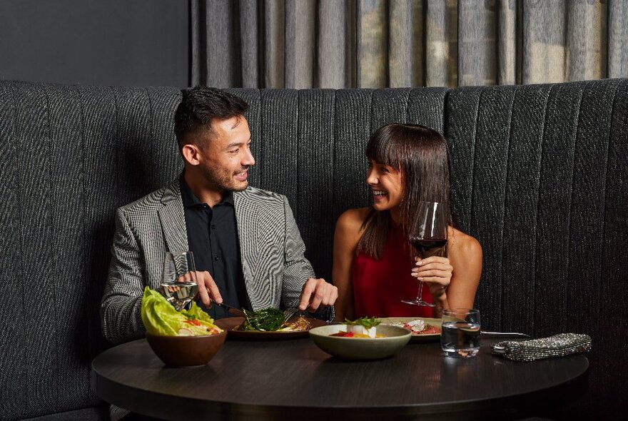A couple seated at a restaurant banquette, eating an array of dishes and drinking red wine.