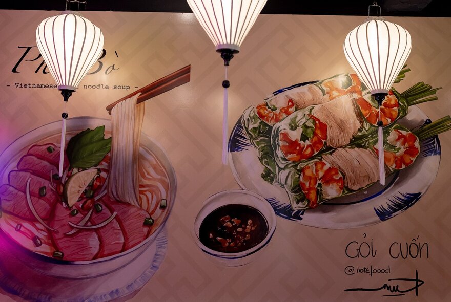 Three paper lanterns in front of a wall-size mural of Pho Bo, Vietnamese beef noodle soup, and Goi Cuon, rice paper rolls.