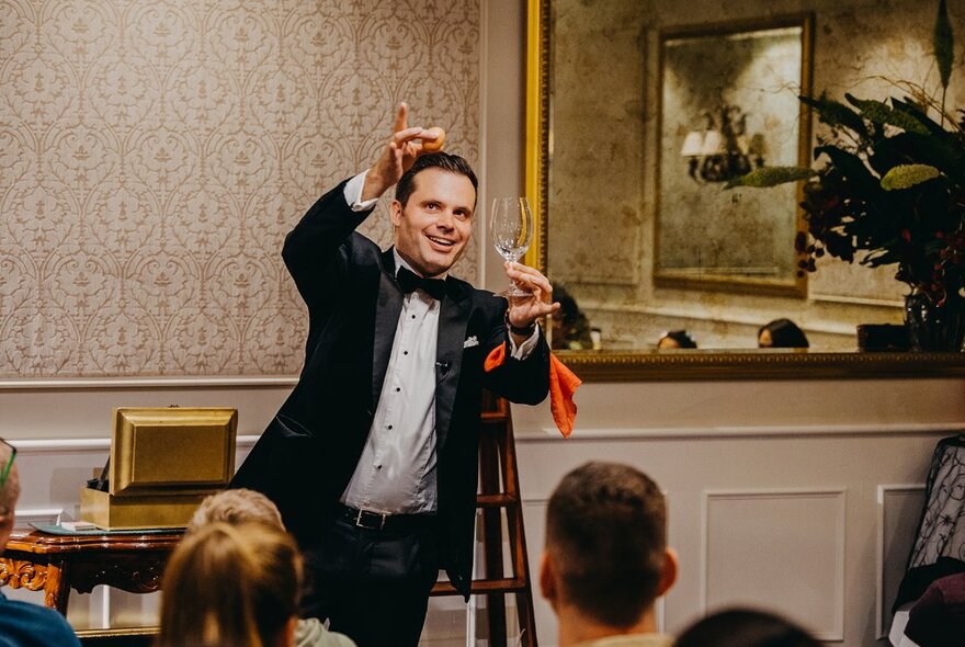 Man wearing a tuxedo performing sleight of hands tricks with a glass in front of a seated audience.