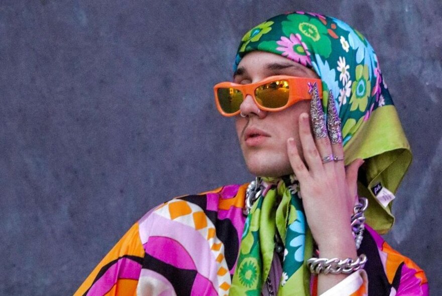 A model poses with a hand on their face in brightly coloured fabrics and a headscarf and orange sunglasses. 