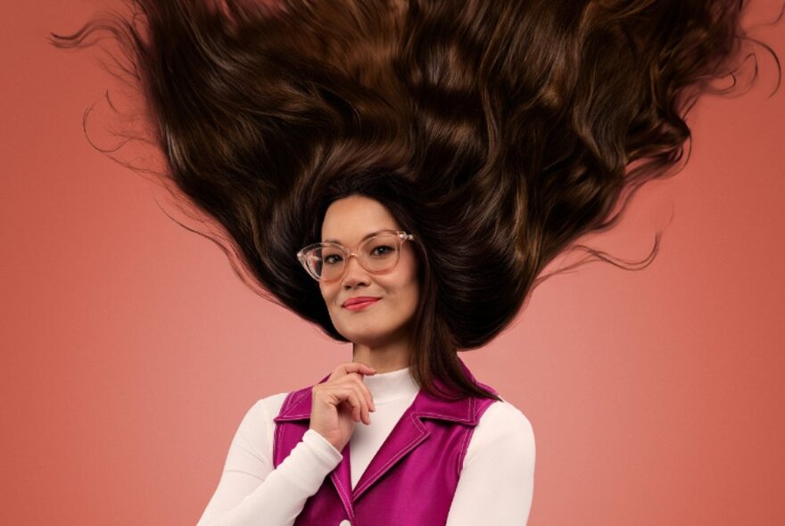 Comedian Lizzy Hoo with her long hair straight up in the air, against a russet pink background.