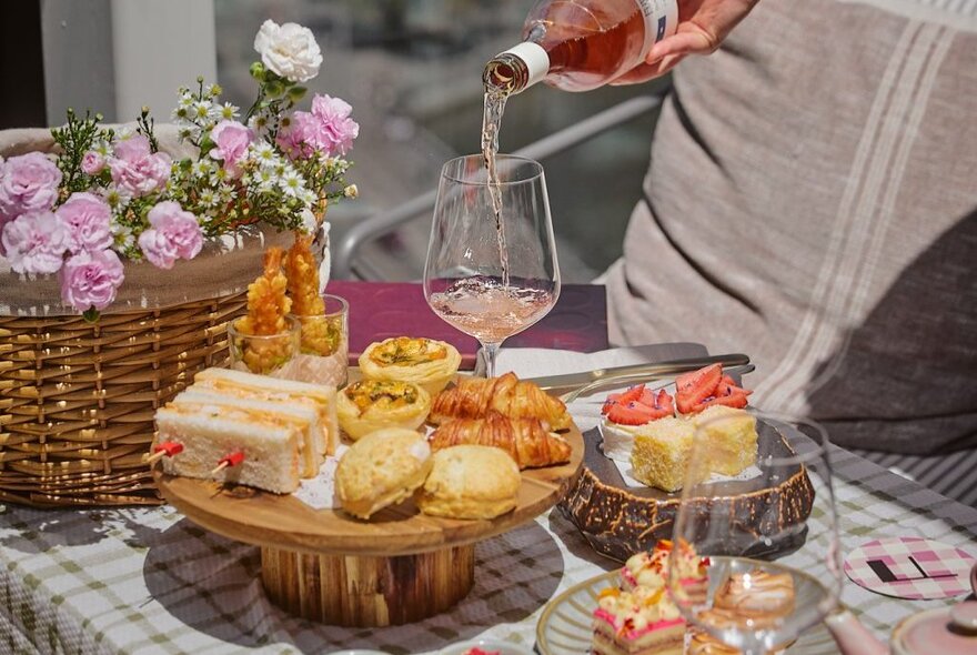 A glass of rose being poured over a high tea picnic set up with sandwiches, pastries and cakes.