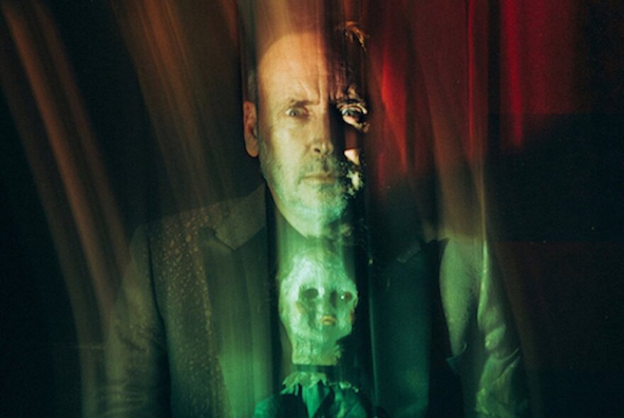 A spooky, blurred image of a man in front of a red curtain, with a creepy dolls head in front of his chest.
