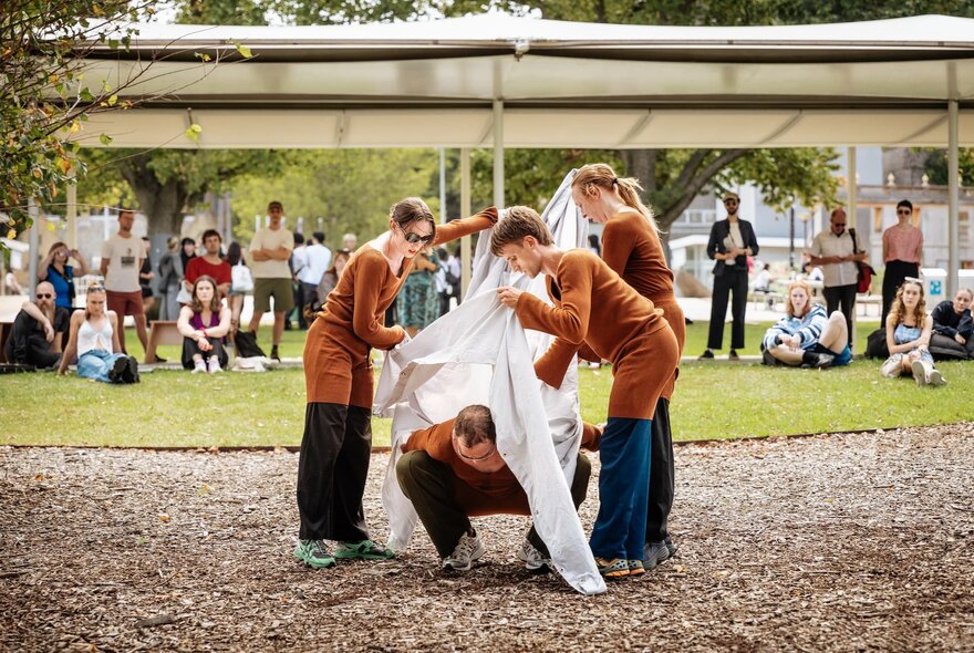 People draping cloth over a person in front of people looking on under a pavilion venue in an outdoor space.