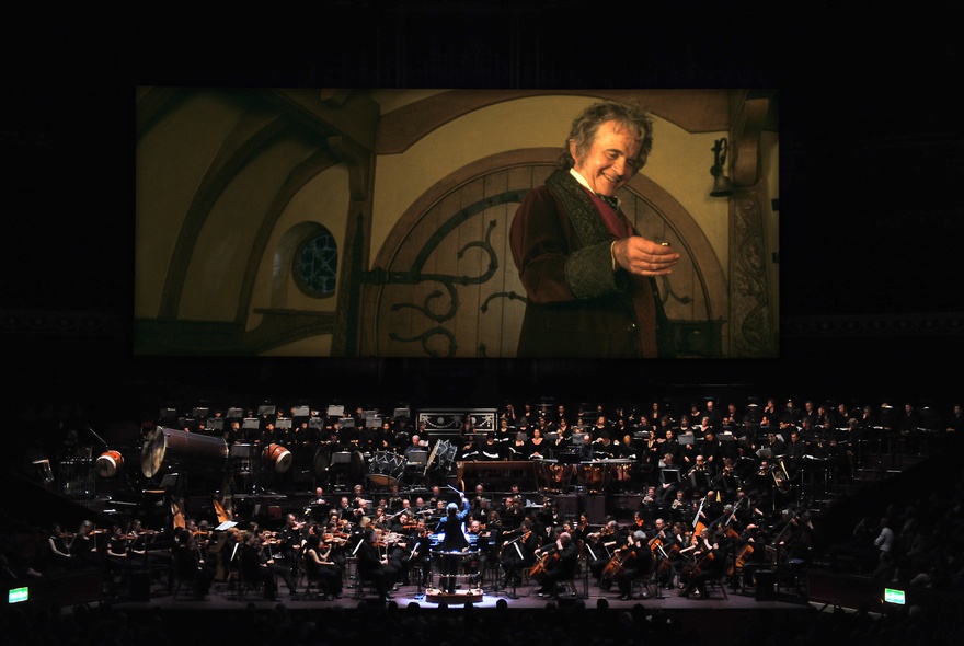 A full orchestra on a stage in front of a large cinema screen showing a still from the movie Lord of the Rings: The Fellowship of the Ring.