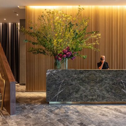 Elegant and spacious foyer of Next Melbourne hotel, showing a large marble check-in counter with an oversize floral arrangement on the desk, and a staircase to the left.