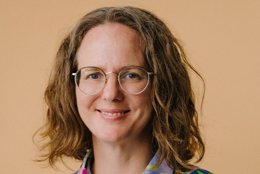 A headshot of a smiling woman with wavy brown hair and round glasses against a tan background.