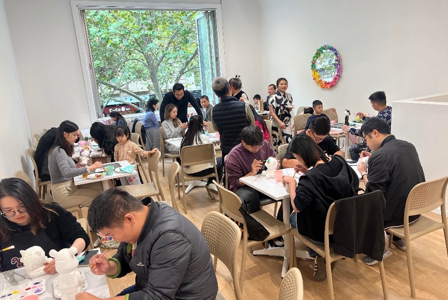 A room full of people seated at tables and painting plaster figurines, with an open window at the back showing a leafy tree.