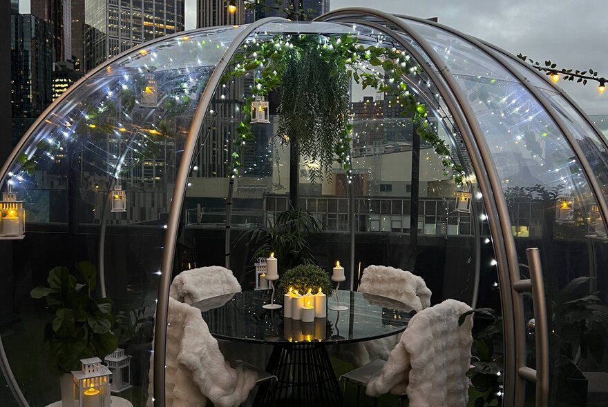 A heated glass dome on a rooftop with a table and four chairs with white throw blankets on them. 