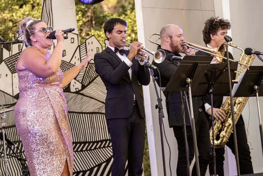 Performer Elly Poletti on stage in a shimmering sequined dress, singing into a microphone alongside three brass instrument players.