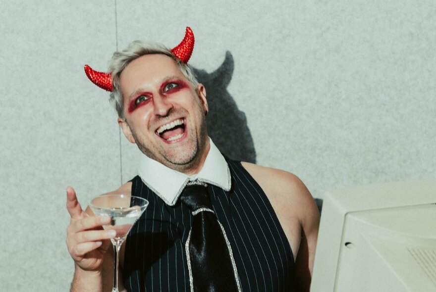 A man dressed as satan laughing and holding up a martini in front of an old computer monitor. 
