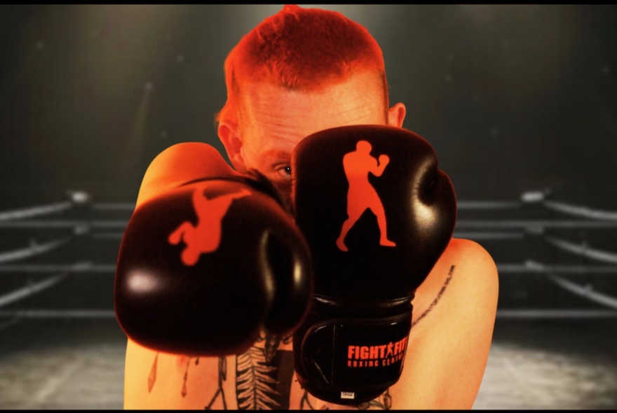 Comedian with short red hair, in a boxing ring, hiding behind boxing gloves.