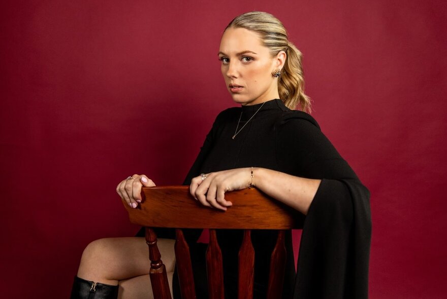 A woman in a drapey black top, seated sideways in a wooden chair in front of a deep red background.