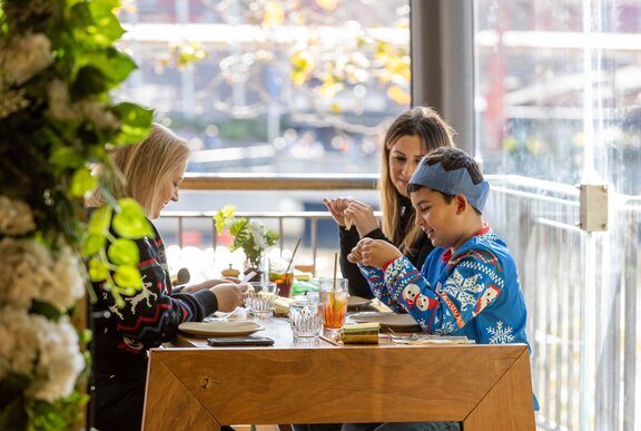 Two women and young boy wearing a paper Christmas hat seated at a restaurant with a view. 