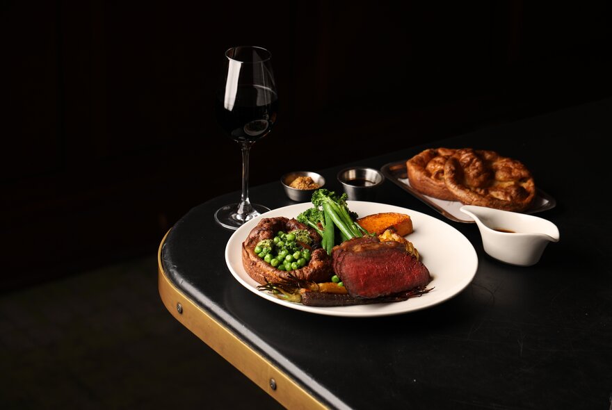 A dark image with a plates of roast meat and vegetables with a glass of red wine. 