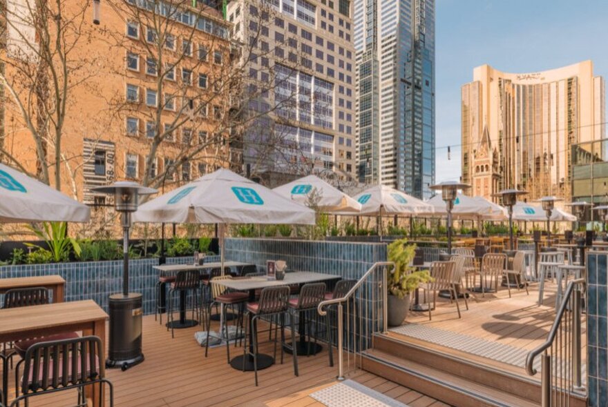 A sunny rooftop bar on tree level in Melbourne's CBD, furnished with large white umbrellas and high wooden tables.