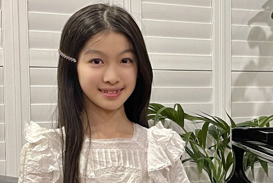 Young pianist, Chloe Au, smiling in a white dress with plantation shutters behind her and a large indoor plant. 