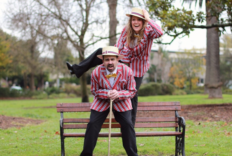 Man and woman outdoors in a park, posing on a park bench, wearing matching striped jackets, straw boaters, and black pants.